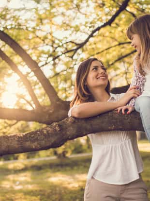 Mother looking her daughter sitting on tree branch at sunny day in public park