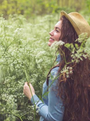 Beautiful, young, longhaired woman with a straw hat and a backpack smiling and standing in the meadow, surrounded by flowers