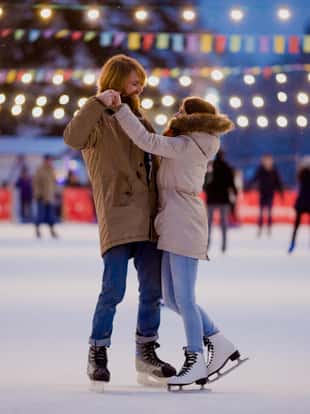 Theme ice skating rink and loving couple. meeting young, stylish people ride by hand in crowd on city skating rink lit by light bulbs and lights. Ice skating in winter for Christmas on ice arena.