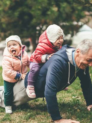 A photo of a playful grandfather and granddaughter. They are casually dressed and playing in the park. They exercise together. A grandfather is exercising while granddaughters are sitting on his back.