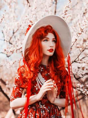 Young handsome redhead girl in spring flower garden. Woman in vintage hat and retro dress on nature. Beautiful spring cherry tree. Handsome redhead model in retro hat. Vanilla flower dress. Red nails