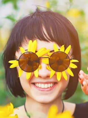 Close up creative portrait of a beautiful young smiling happy brunette girl with yellow flower petals under sunglasses on background of a field of sunflowers. Woman summer lifestyle and healthy teeth