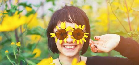 Close up creative portrait of a beautiful young smiling happy brunette girl with yellow flower petals under sunglasses on background of a field of sunflowers. Woman summer lifestyle and healthy teeth