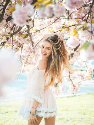 Girl in short pink dress enjoying sunny day in botanical garden. Female playing with long gorgeous blond hair, beauty and hair care products. Sexy woman posing under blooming cherry blossom tree.