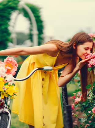 a cute and stylish girl in a yellow dress and long hair rides on a bicycle with a basket of flowers in the sunny summer garden