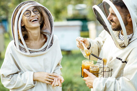 Portrait of a cheerful man and woman beekepers in protective uniform standing together with honey in the jar, tasting fresh product on the apiary outdoors