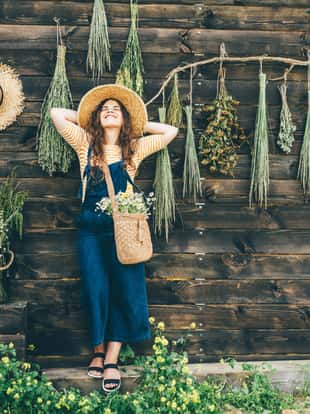 Attractive girl wearing straw hat and blue denim dungarees relaxing near bouquets of aromatic wildflowers on wooden old summerhouse wall on sunny day.