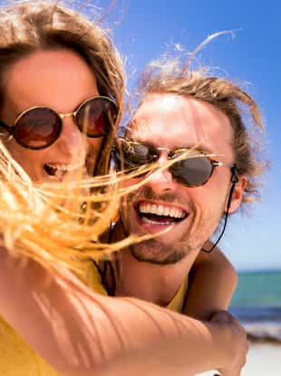 Close-up of cheerful boyfriend piggybacking girlfriend against sky. Happy couple is enjoying at beach during summer. They are wearing sunglasses on sunny day.