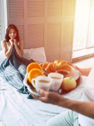 Handsome young man is standing with romantic breakfast on tray, he made it for his beautiful smiling cute girl or wife, who is lying on bed and waiting for him