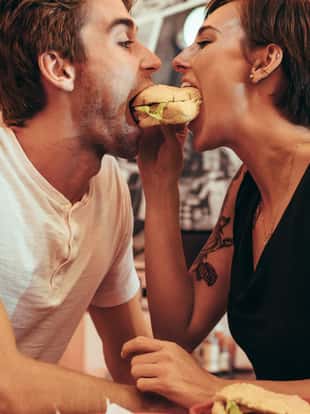 Romantic couple eating a burger together at a restaurant looking at each other. Couple dining at a restaurant biting a burger together.