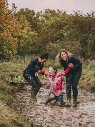 Women are taking part in a charity obstacle course. One woman is stuck in a muddy ditch and the other two are trying to help her get out. They are laughing to hard to be able to lift her.