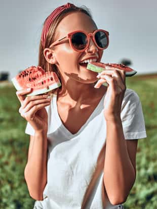 Sexy girl in red sunglasses holds a cut watermelon in her hands. Happy woman in trendy red sunglasses eat watermelon on nature
