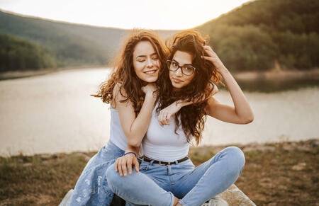 Two beautiful young female friends sitting on lakeshore, enjoying summer sunset by the lake and hills