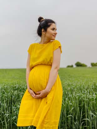 Photo of a pregnant woman standing alone on a grass field, stroking her baby bump and enjoying the calm meadow.