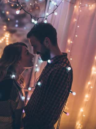 Young couple is kissing by the frosted window decorated with Christmas lights, while it is snowing outside