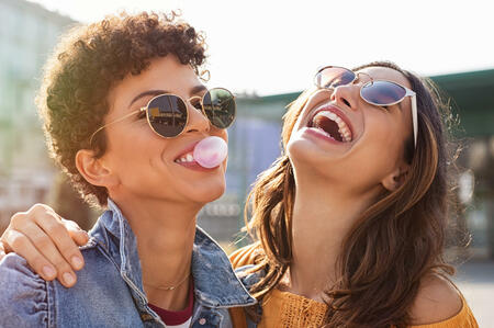 Young latin woman laughing while friend inflating bubble gum. Closeup face of multiethnic friends enjoying outdoor street. Brazilian girl laughing and blowing chewing gum with friend embracing and looking up.