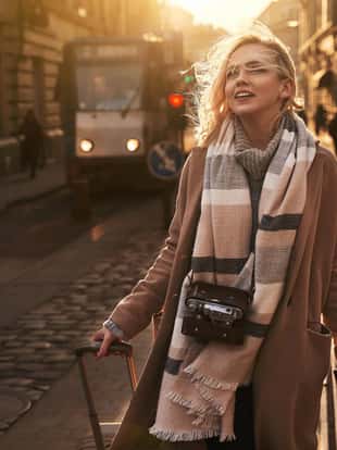 Young beautiful blond woman tourist with a wheeled travel bag and vintage film camera arrives to a new city on a sunny windy evening at golden hour.