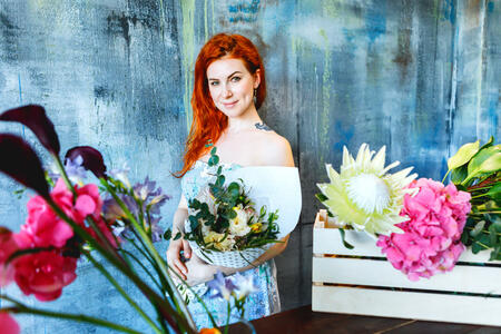 Charming cheerful red hair female shop assistant stands by wooden box with flowers with Hydrangea purple, freesia, protea, roses. Flower shop and floristic design master class concept on painted blue wall