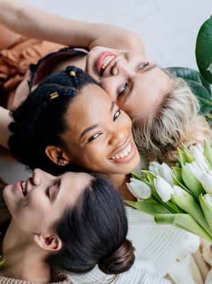 Group of three happy multiethnic sits together on floor. Portrait of women of different cultures enjoying bouquet of tulips. Smiling blond girl with two african american friends. Spring mood