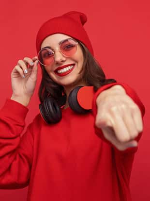 Stylish smiling brunette in red sunglasses and sweatshirt pointing happily at camera making choice on red backdrop