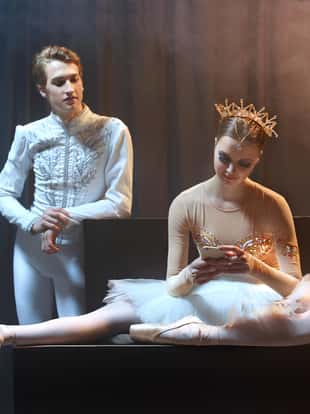 Classical ballet dancers have a rest after performing on stage in theatre. Woman is looking into the phone, while a man is standing from behind.
