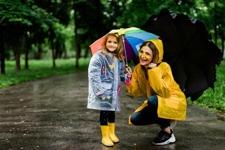 Happy mother and daughter holding umbrellas and wearing rubber boots while walking