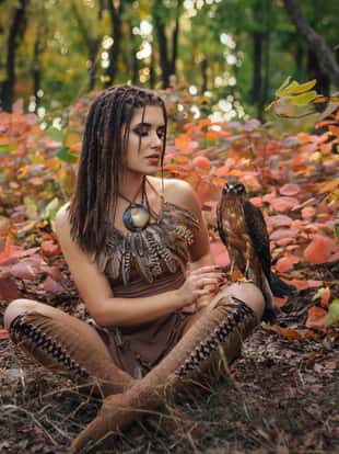 Aggressive-sexual wild girl, wanders in the jungle with a tamed bird. Princess warrior in a brown dress and a necklace of feathers. Background autumn landscape. Hairstyle dreadlocks. Artistic Photography