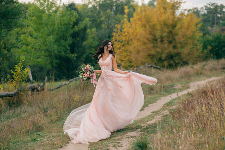 The brunette girl with long hair is happily danced in a pink dress with a train. A bride with a bouquet is running along the path. Film photo.
