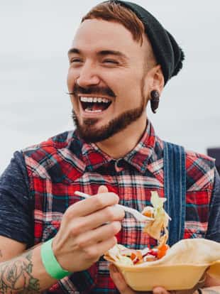 Young male hipster with tattoos is at a music festival. He is eating a teakaway doner kebab while laughing and looking out the frame.