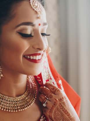 Stunning Indian bride dressed in Hindu red traditional wedding clothes lehenga embroidered with gold and a veil smiles tender