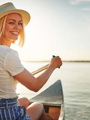 Smiling young blonde woman looking back over her shoulder while paddling a canoe on a scenic lake on a summer afternoon