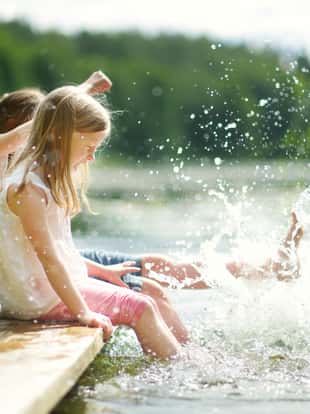 Two cute little girls sitting on a wooden platform by the river or lake dipping their feet in the water on warm summer day. Family activities in summer.