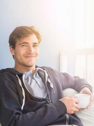 Portrait of young man holding coffee cup while sitting on sofa. Handsome male is in casuals. He is smiling at home.