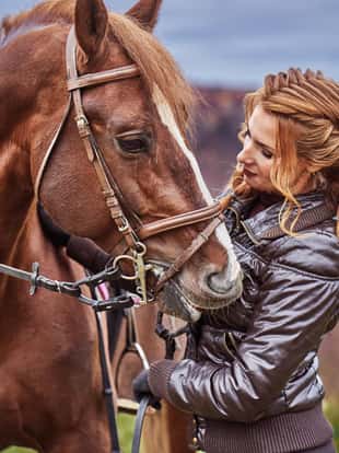 A young woman with brown curly hair caresses a brown horse. The horse caresses the woman. Woman on a walk with a horse on a cloudy autumn day.