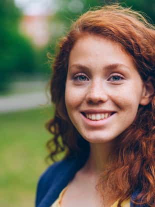 Close-up portrait of beautiful young lady with long red hair and freckles smiling looking at camera in park in summer. Nature, people and happiness concept.