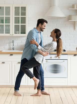 Happy romantic young couple bonding dancing in modern kitchen with white furniture enjoy sweet moments of affection, cheerful husband and wife celebrating anniversary having fun at home lifestyle