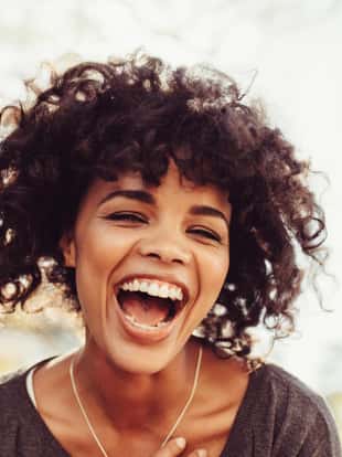 Close up of young woman with curly hair laughing. Woman standing in morning sun in cheerful mood.