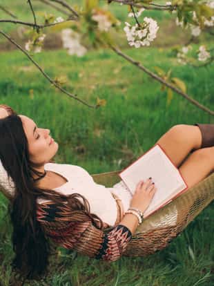 Pretty young brunette woman is reading book in hammock in blossom cherry garden with white flowers and green grass. Spring time.