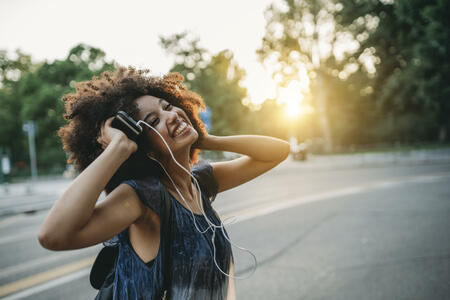 Young adult woman with afro hair dancing in the city at sunset while listening to music