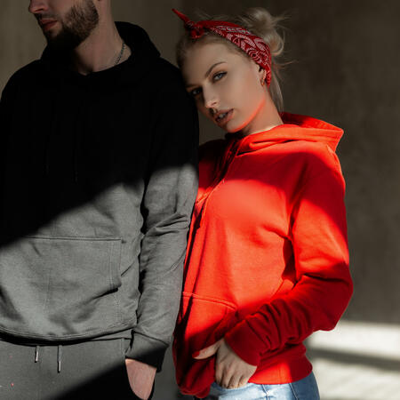 Beautiful young stylish couple in trendy hoodie indoors with sunshine. Hipster girl in red hoodie and handsome man with beard in blue hoodie in studio